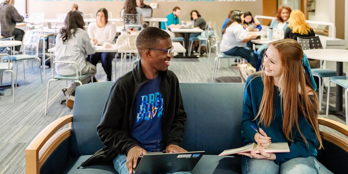 Two students, one on a laptop, one writing in a notebook, talk in the Testing Center Lounge