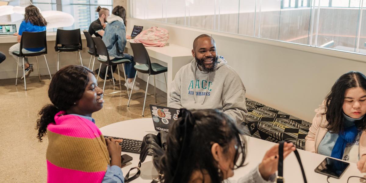 A group of students sits at  table talking. Some are working on computers.